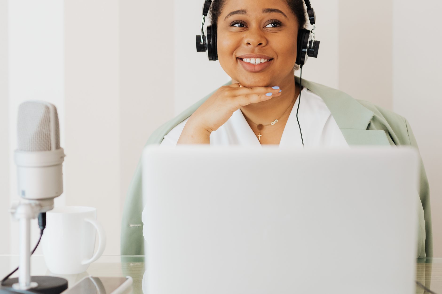 Mujer sonriente con auriculares y un micrófono de mesa, sentada detrás de un portátil en un espacio luminoso.