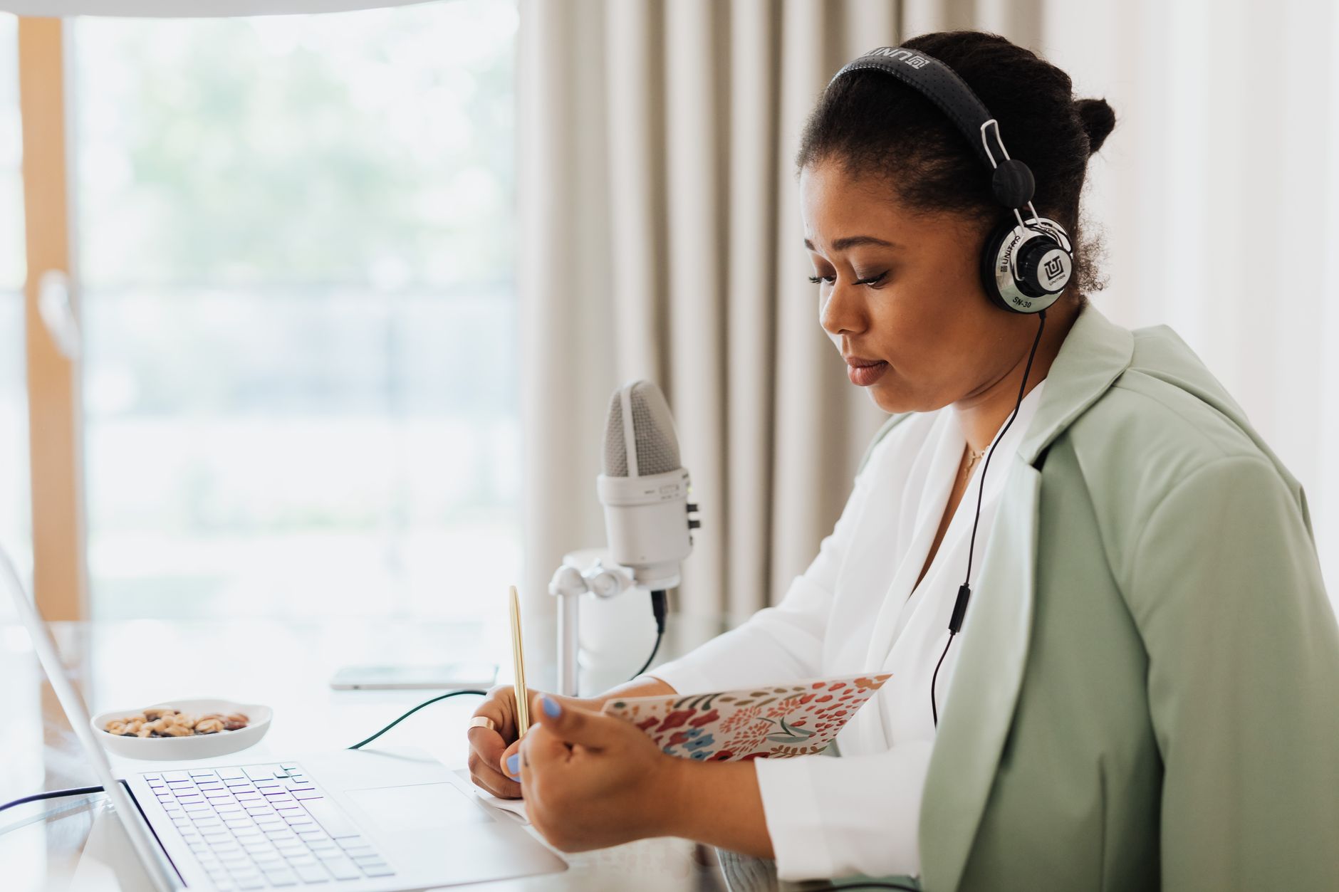 Mujer con auriculares tomando notas junto a un portátil y un micrófono, cerca de una ventana.