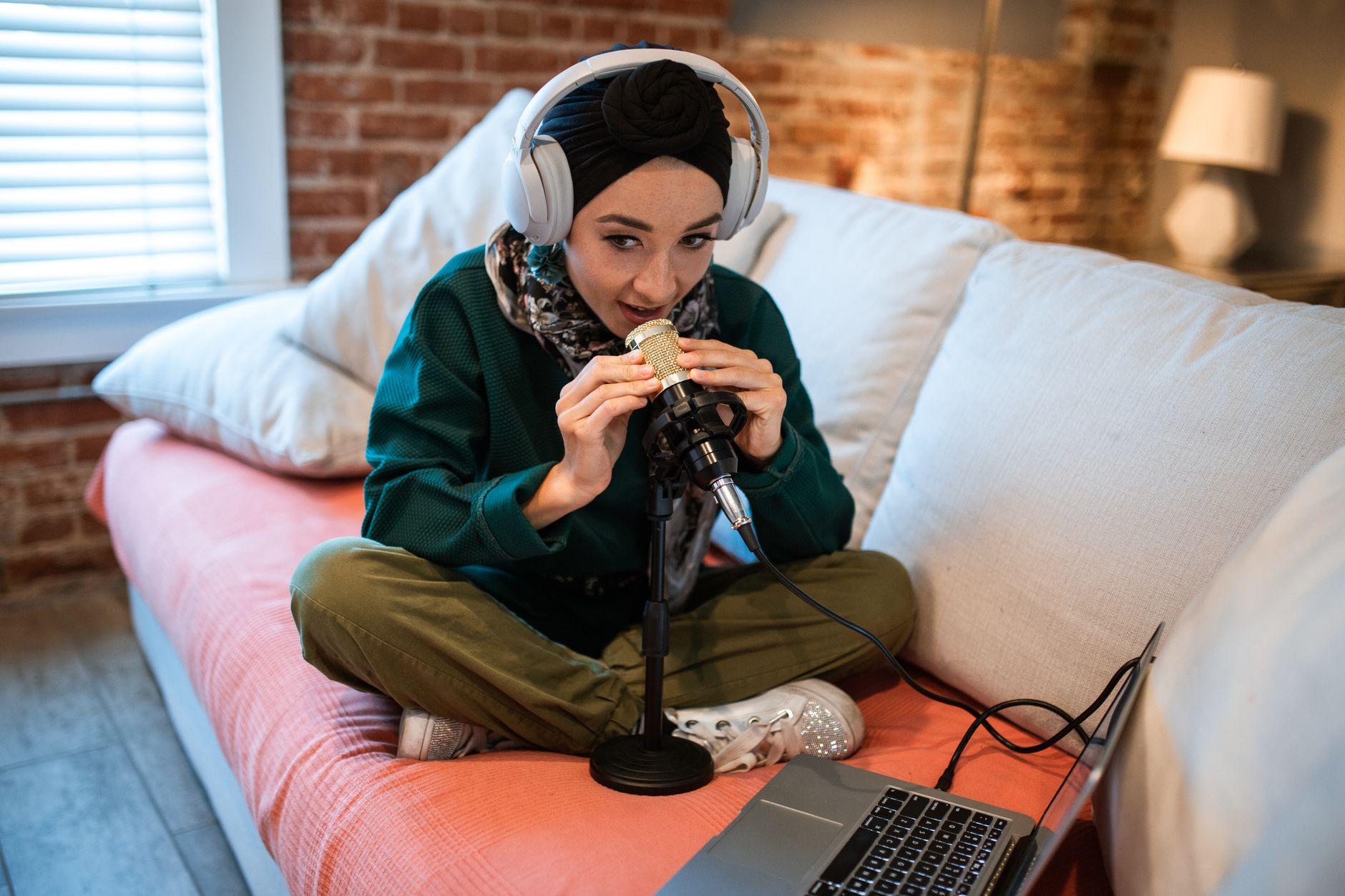 Mujer con auriculares sentada en un sofá, hablando ante un micrófono con un portátil delante.