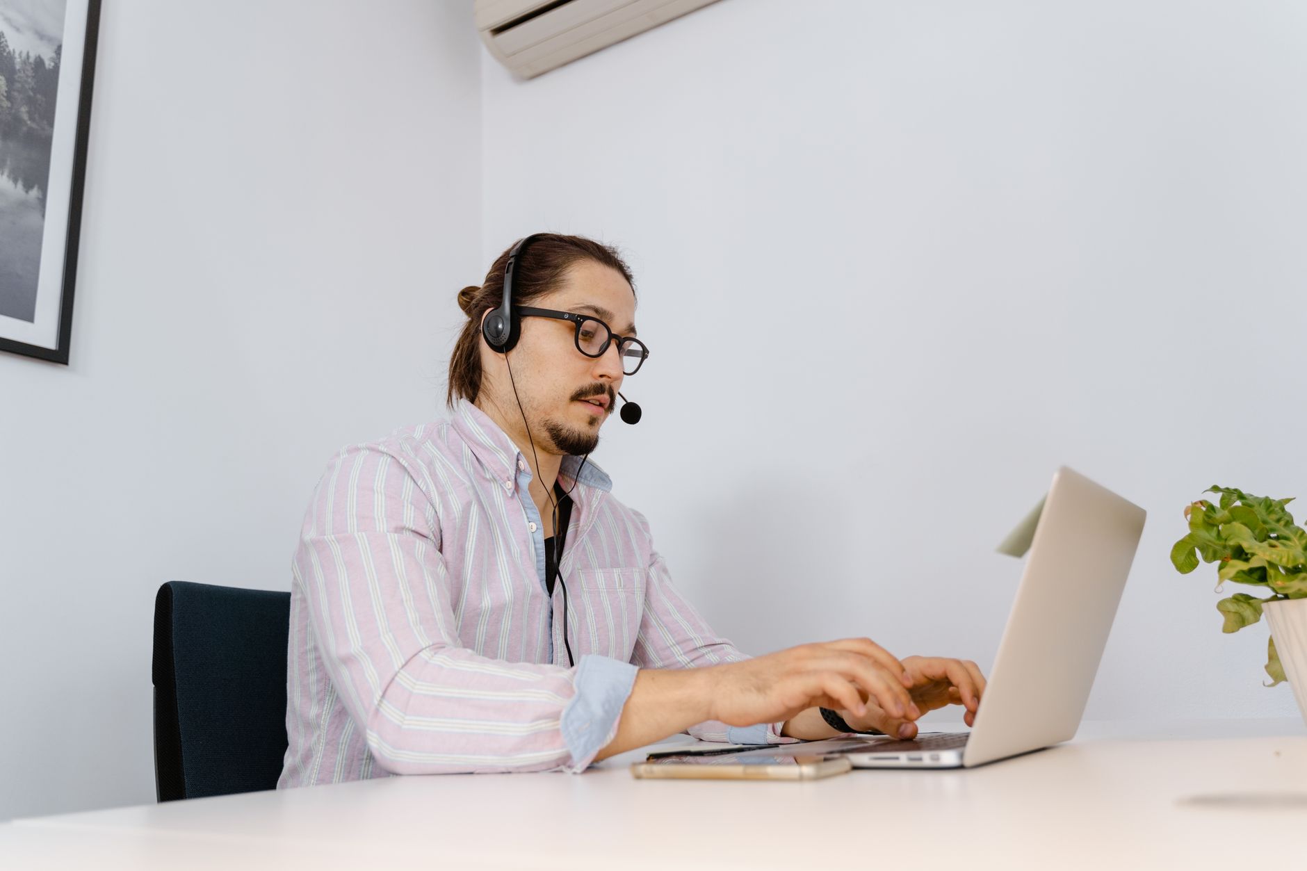 Hombre con gafas y auriculares con micrófono escribiendo en un portátil sobre una mesa blanca.